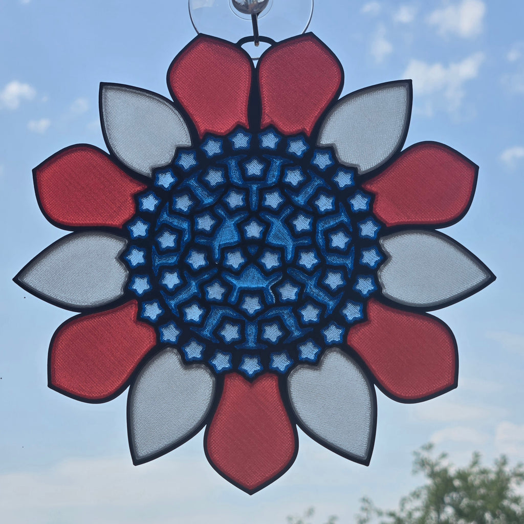 A circular 3D printed sun catcher with a red, white, and blue floral design, suspended by a metal ring against a blue sky background.