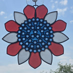 A circular 3D printed sun catcher with a red, white, and blue floral design, suspended by a metal ring against a blue sky background.