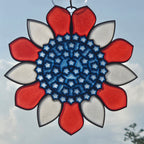 A 3D printed sunburst ornament with a blue center and alternating red and white outer petals, suspended against a cloudy sky.