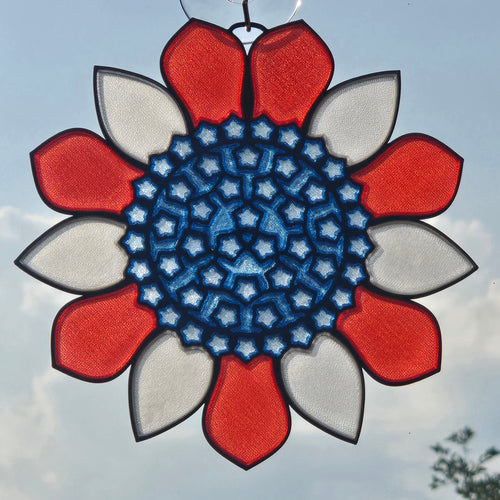 A 3D printed sunburst ornament with a blue center and alternating red and white outer petals, suspended against a cloudy sky.