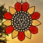A 3D printed sunflower ornament hanging in warm backlighting, with red and gold glass petals, a dark center, and small star-shaped details.