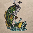 A 3d printed style fish ornament or wall hanging with a green and white fish jumping over blue seaweed, suspended from a clear hook against a tan background.