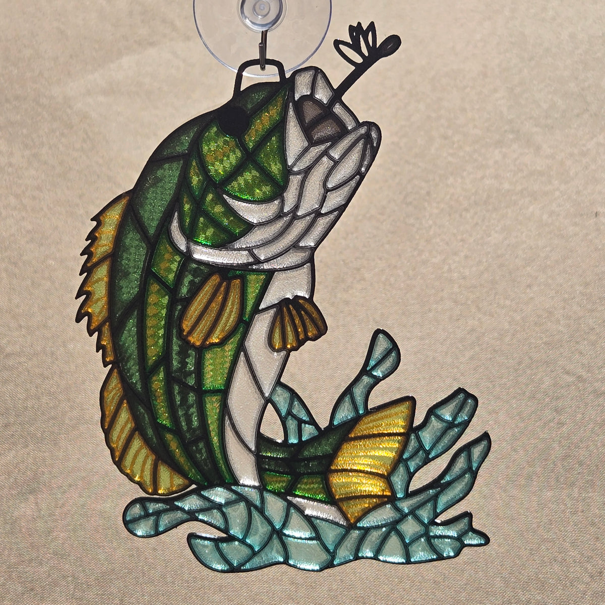 A 3d printed style fish ornament or wall hanging with a green and white fish jumping over blue seaweed, suspended from a clear hook against a tan background.
