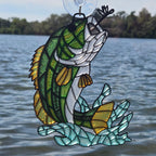 A 3d printed bass fish ornament with green, yellow, white, and blue pieces, attached to a clear suction cup and photographed outdoors over water with trees in the background.