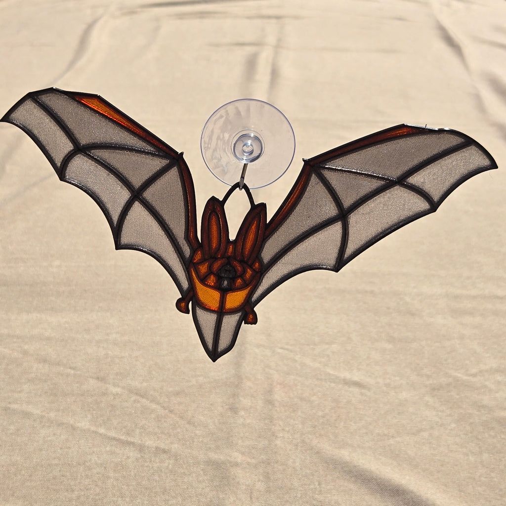 A 3D printed bat ornament with translucent gray wings, black outlines, and orange accents, attached to a clear suction cup against a beige fabric background.