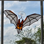 A bat-shaped 3D printed suncatcher with orange and amber panels, white wings, and a clear suction cup at the top, hanging in front of a window.