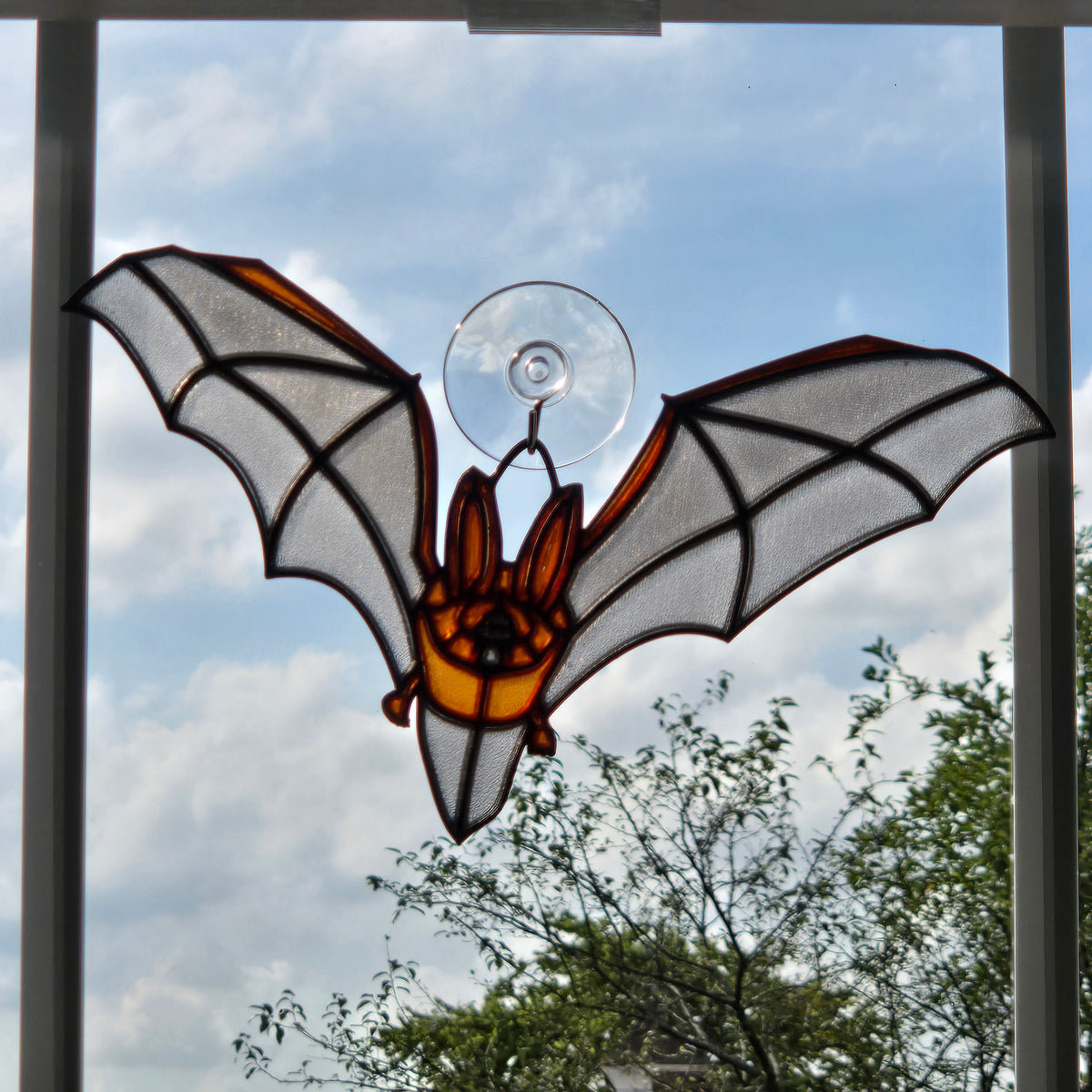 A bat-shaped 3D printed suncatcher with orange and amber panels, white wings, and a clear suction cup at the top, hanging in front of a window.