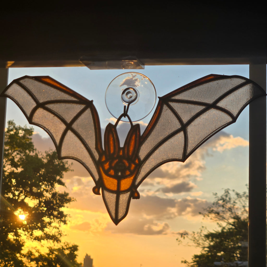 A 3D printed bat suncatcher hanging from a clear suction cup on a window, backlit by a warm sunset sky with trees visible in the background.