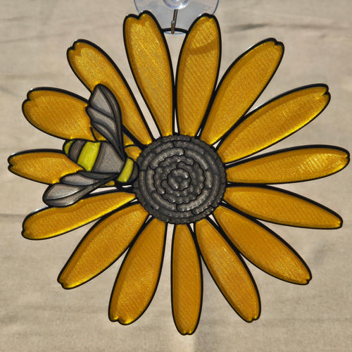 A 3D printed black eyed susan ornament with a bumblebee on one of its yellow petals, suspended by a clear plastic hook against a neutral background.