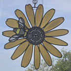 A 3D printed suncatcher shaped like a black eyed susan with a bee on the left petal, suspended by a clear hook against a blue sky background with blurred green trees.