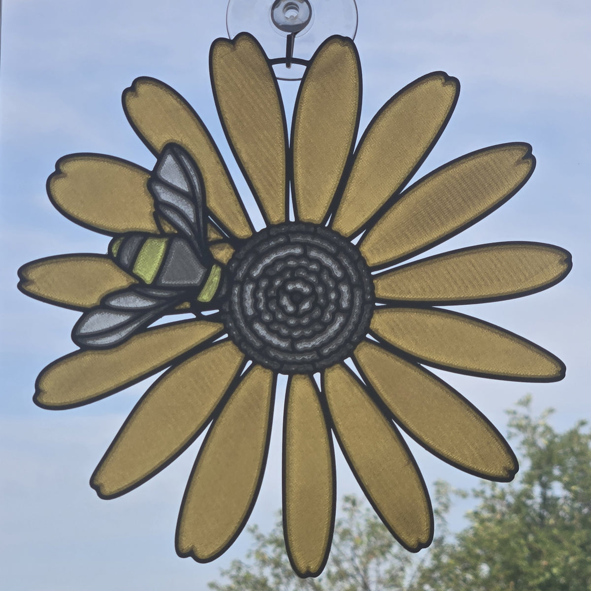 A 3D printed suncatcher shaped like a black eyed susan with a bee on the left petal, suspended by a clear hook against a blue sky background with blurred green trees.