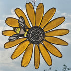 A 3D printed suncatcher shaped like a black eyed susan with a bee on the left side, suspended by a metal wire against a blue sky background.