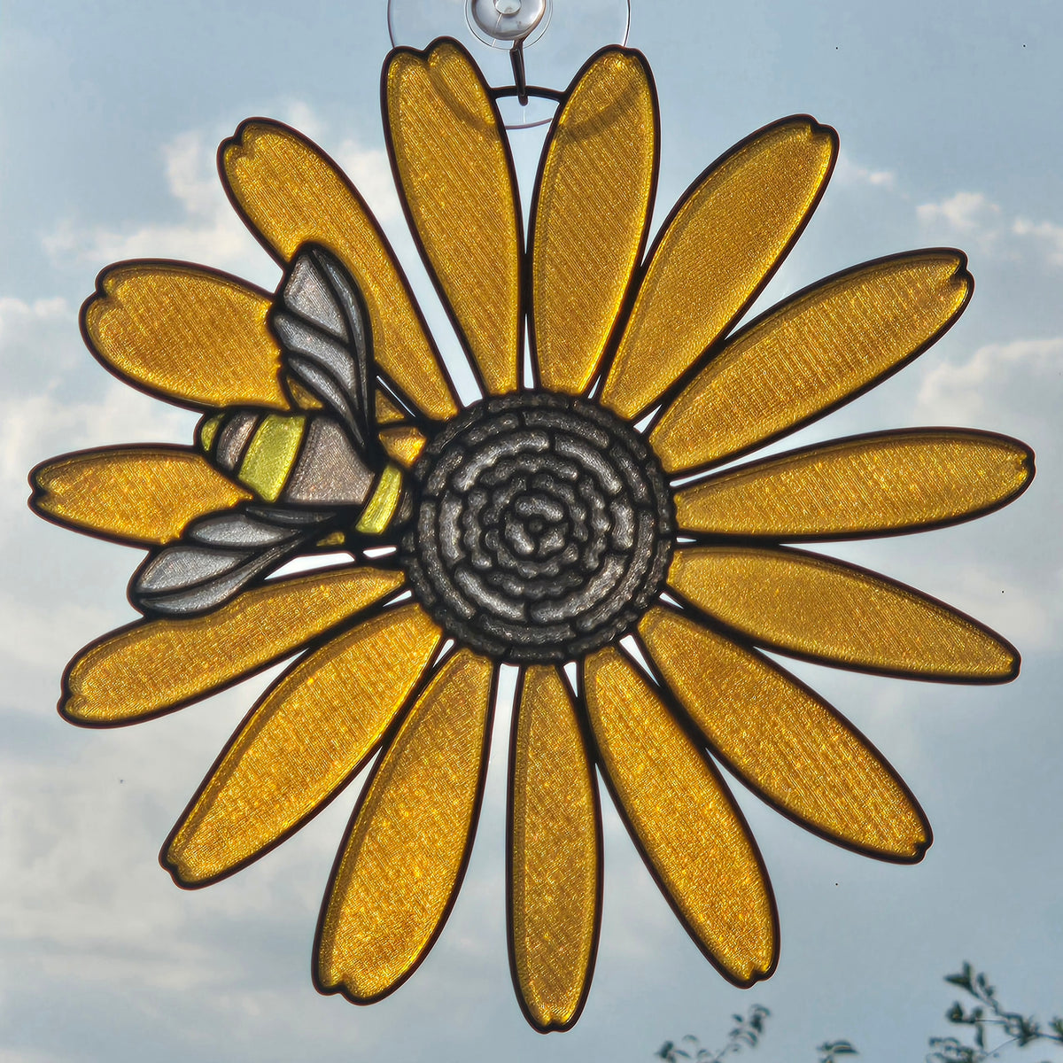 A 3D printed suncatcher shaped like a black eyed susan with a bee on the left side, suspended by a metal wire against a blue sky background.