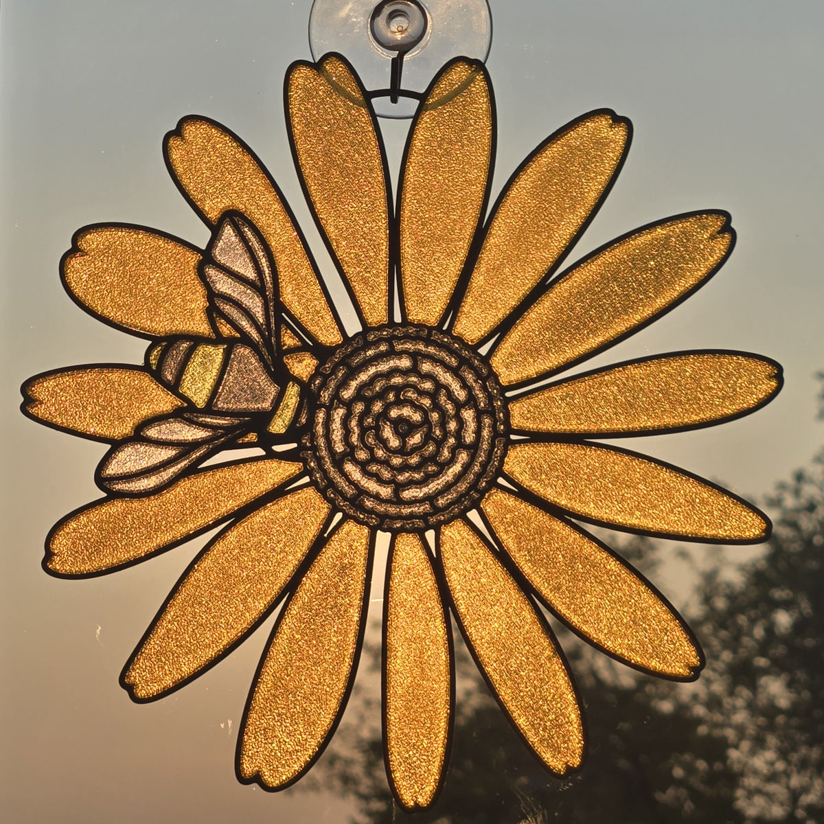 A 3D printed hanging ornament shaped like a yellow flower with a bee on the left side, suspended by a clear hook against a softly lit outdoor background.
