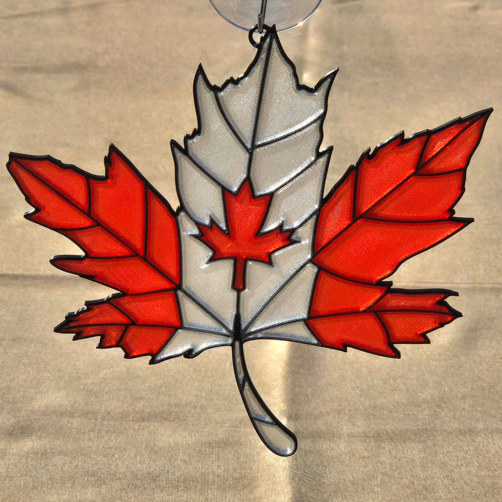 A 3D printed ornament shaped like a Canadian maple leaf, featuring red and white glass panels outlined in black metal, suspended from a clear hanging loop against a beige fabric background.