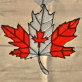 A 3D printed ornament shaped like a Canadian maple leaf, featuring red and white glass panels outlined in black metal, suspended from a clear hanging loop against a beige fabric background.