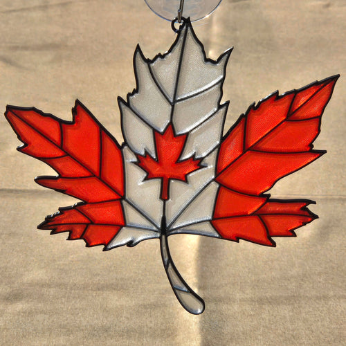 A 3D printed ornament shaped like a Canadian maple leaf, featuring red and white glass panels outlined in black metal, suspended from a clear hanging loop against a beige fabric background.