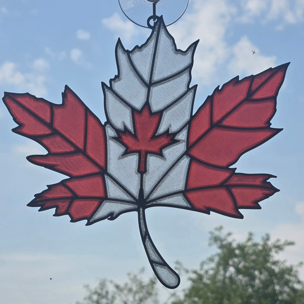 A 3D printed Canadian maple leaf ornament hanging in front of a blue sky with clouds and green trees in the background.