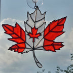 A 3D printed maple leaf ornament featuring red and white sections with black leading, suspended by a clear suction cup against a cloudy blue sky.