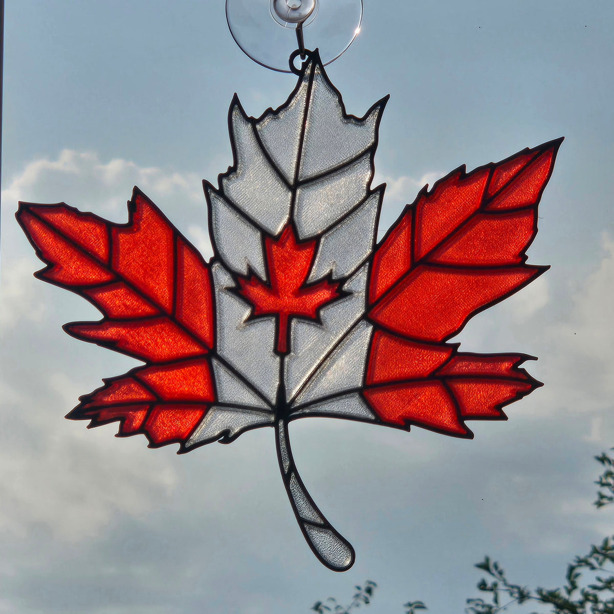 A 3D printed maple leaf ornament featuring red and white sections with black leading, suspended by a clear suction cup against a cloudy blue sky.