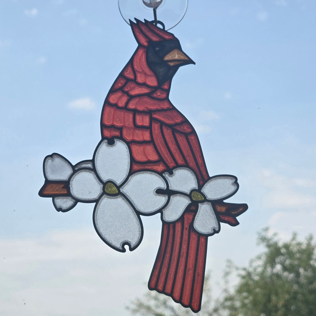 A 3D printed suncatcher depicting a cardinal perched on white dogwood flowers, suspended against a blue sky background.