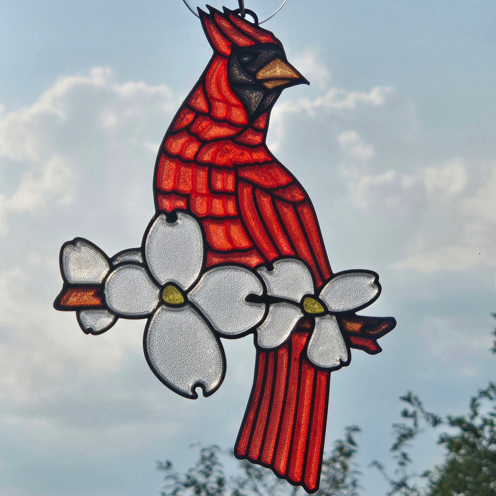 A 3D printed suncatcher depicting a cardinal perched on a flowering branch, suspended against a cloudy sky background.