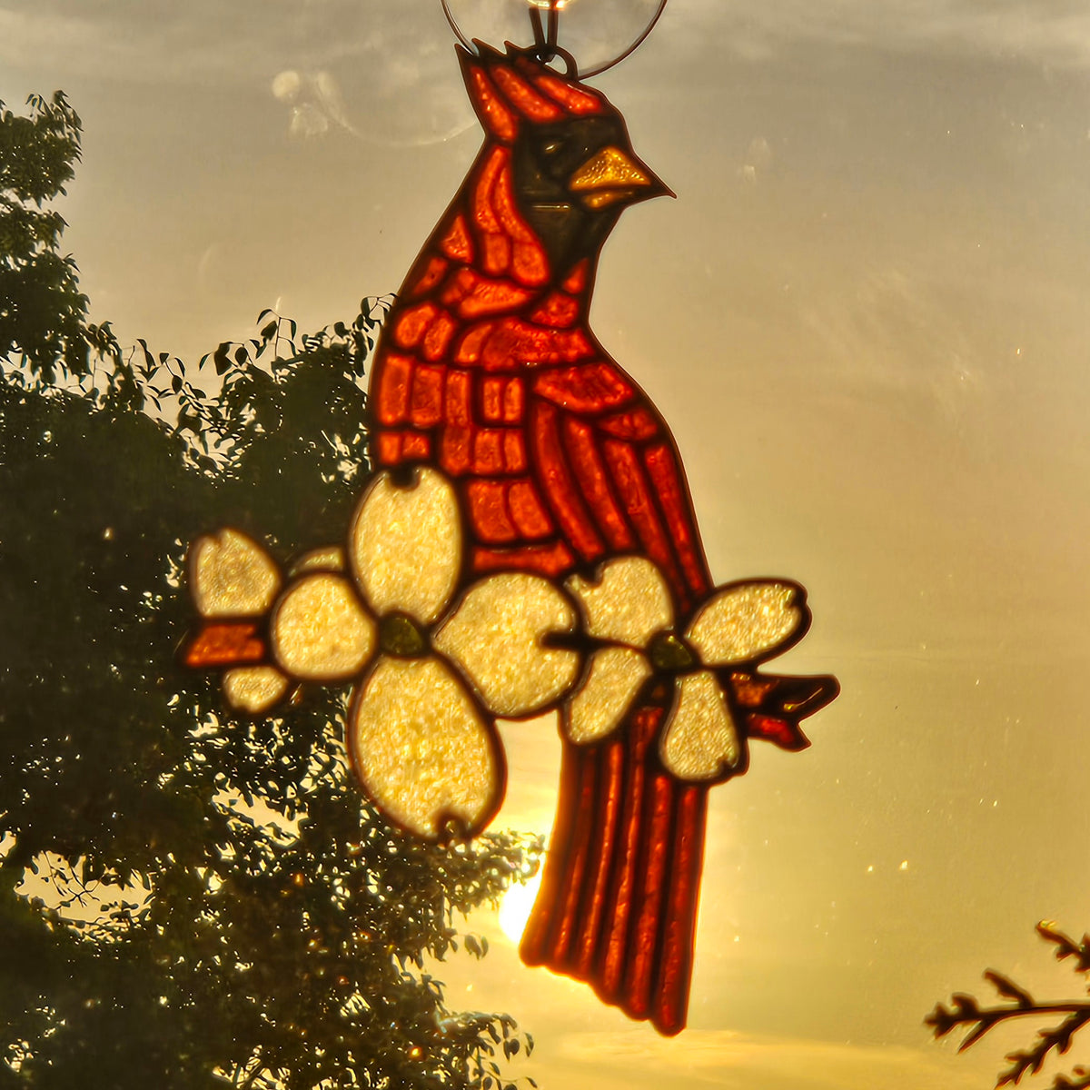 A 3D printed suncatcher shaped like a cardinal perched on a flowering branch, shown hanging in front of a warm sunset sky with tree silhouettes.