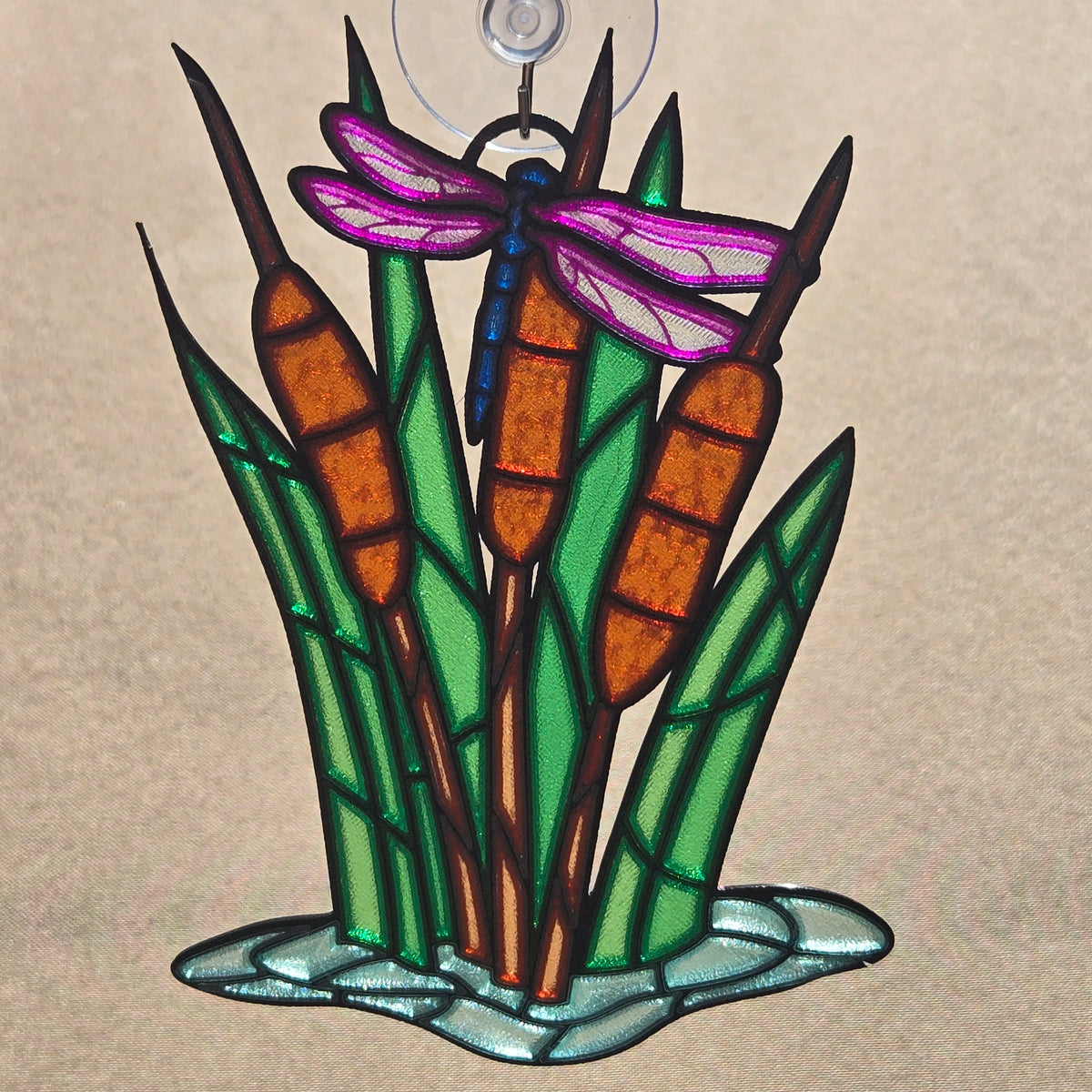 A 3d printed ornament depicting cattails, reeds, and a dragonfly, with a hanging loop at the top, shown against a neutral beige background.