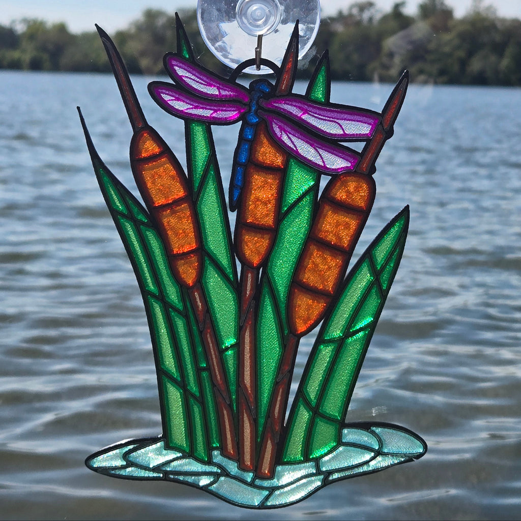 A 3d printed decorative window panel featuring cattails, green reeds, a purple dragonfly, and a clear suction-cup top, shown against a real outdoor lake background.