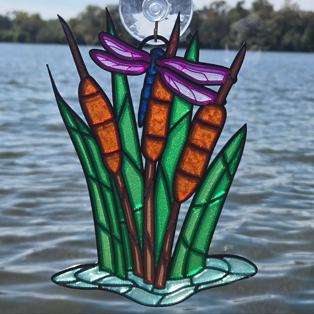 A 3d printed decorative window panel featuring cattails, green reeds, a purple dragonfly, and a clear suction-cup top, shown against a real outdoor lake background.