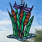 A 3d printed hanging ornament depicting cattails with a dragonfly, suspended by a clear hook against a blue sky background.