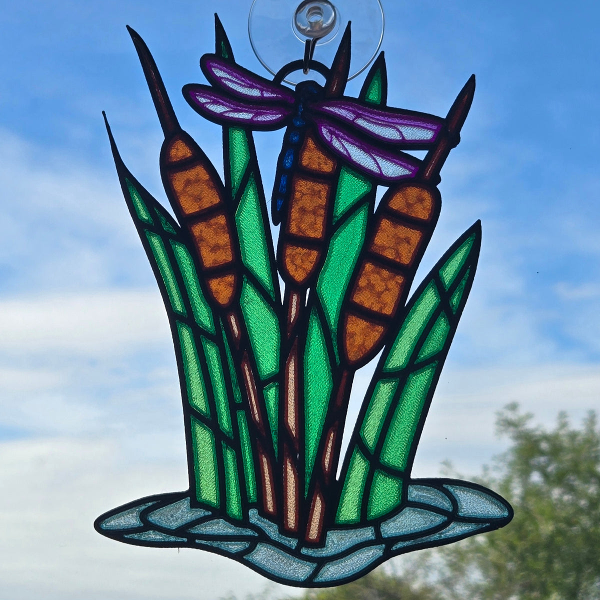 A 3d printed hanging ornament depicting cattails with a dragonfly, suspended by a clear hook against a blue sky background.