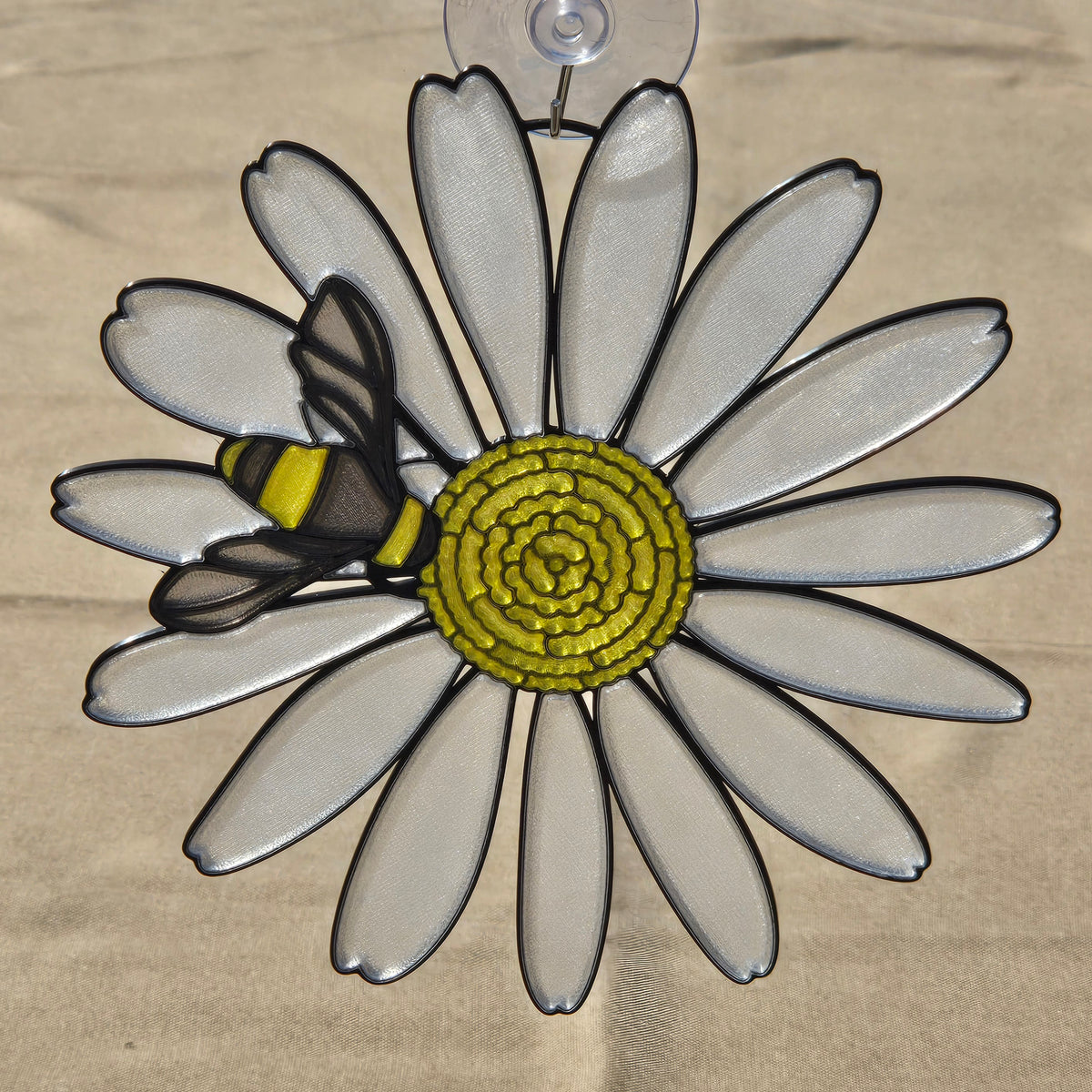 A 3D printed ornament shaped like a daisy with a bee on it, hanging from a clear suction cup against a neutral background.