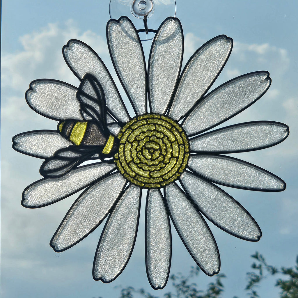 A 3D printed suncatcher shaped like a daisy with a bee on it, hanging against a blue sky background.