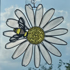A 3D printed suncatcher shaped like a daisy with a bee on it, hanging against a blue sky background.