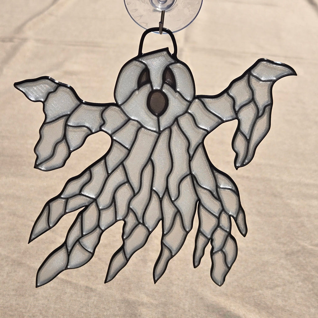 A ghost-shaped 3D printed ornament with a clear suction cup at the top, featuring a white and gray patterned body outlined in black, set against a beige fabric background.