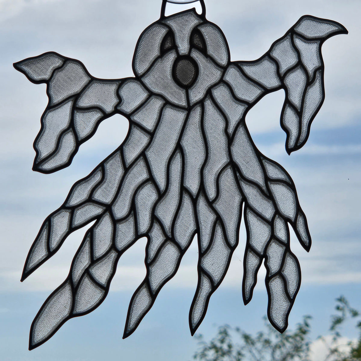 A ghost-shaped 3D printed window ornament with a black metal hanger at the top, shown hanging against a cloudy blue sky background.