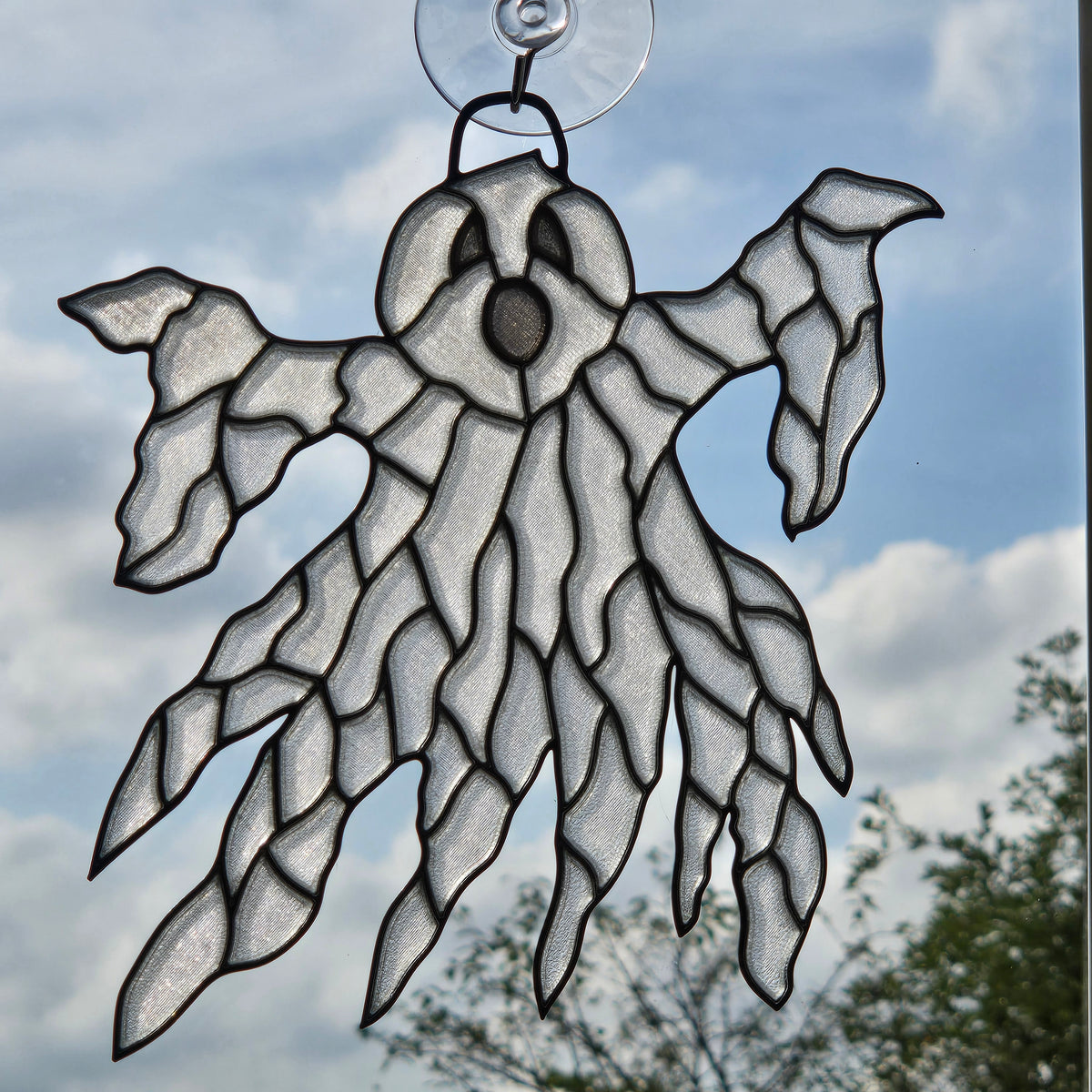 A ghost-shaped 3D printed suncatcher with a clear suction cup at the top, shown hanging against a blue sky with clouds and trees in the background.