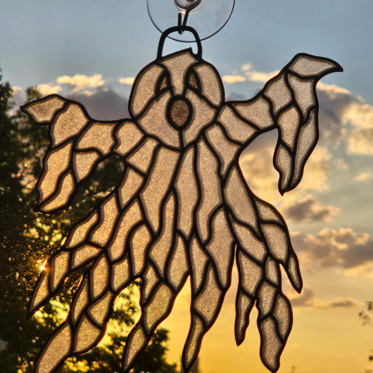A ghost-shaped 3D printed suncatcher with black metal outlines and a clear glittery inner panel, shown hanging in front of a sunset sky with clouds and tree silhouettes.