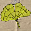 A ginkgo leaf-shaped suncatcher with a translucent yellow-green mesh body and black metal frame, attached to a clear suction cup, shown against a beige cardboard background.