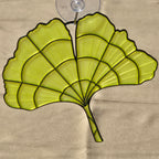 A ginkgo leaf-shaped suncatcher with a translucent yellow-green mesh body and black metal frame, attached to a clear suction cup, shown against a beige cardboard background.