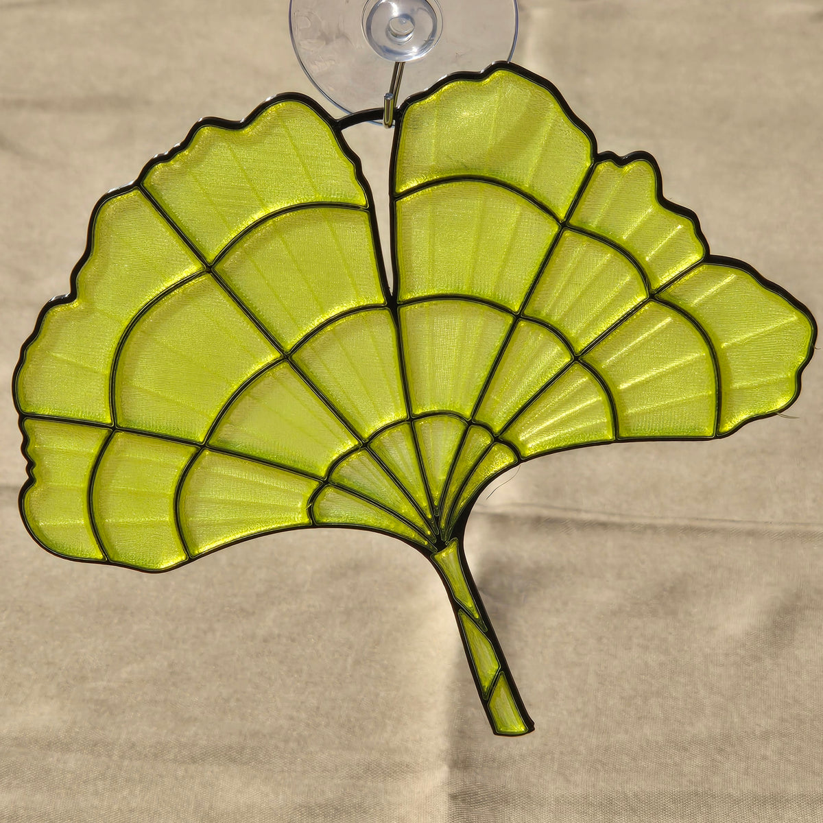 A ginkgo leaf-shaped suncatcher with a translucent yellow-green mesh body and black metal frame, attached to a clear suction cup, shown against a beige cardboard background.