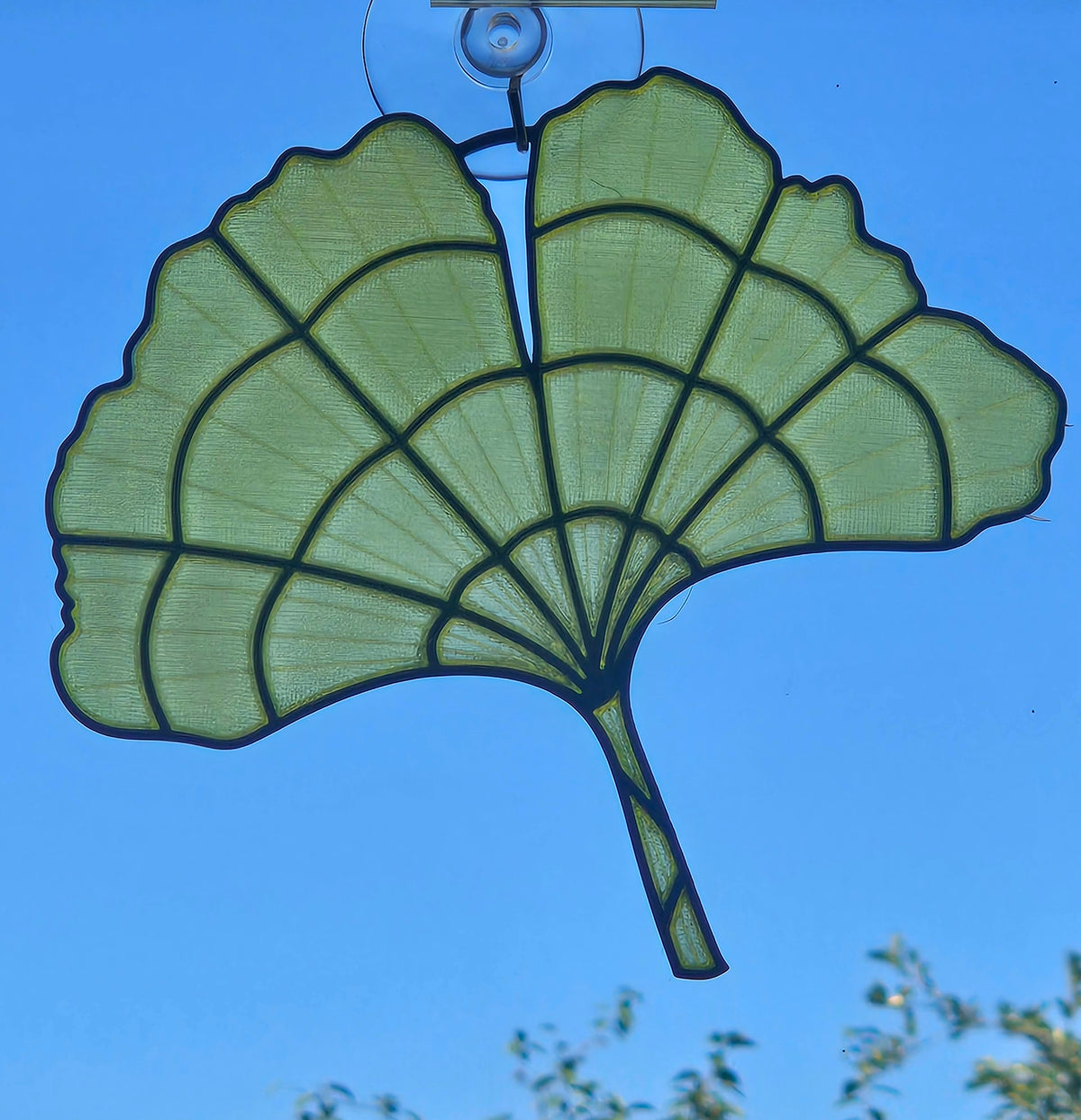 A green ginkgo leaf-shaped hanging ornament with black metal framing, suspended by a clear plastic hook against a bright blue sky.