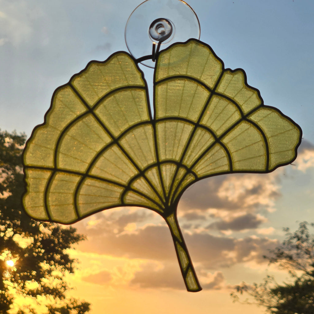 A translucent fan-shaped ginkgo leaf hanging ornament with a metal stem and suction cup, photographed against a sunset sky with clouds and tree silhouettes visible through the glass.