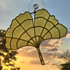 A translucent fan-shaped ginkgo leaf hanging ornament with a metal stem and suction cup, photographed against a sunset sky with clouds and tree silhouettes visible through the glass.