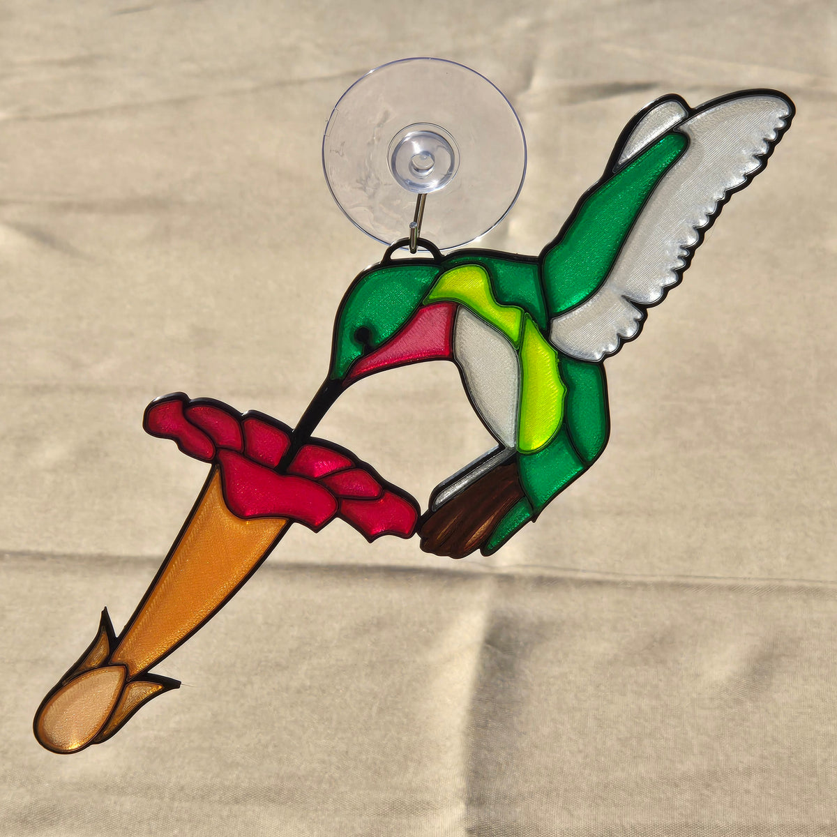 A 3D printed hummingbird ornament featuring a green, yellow, and white bird with a red beak, perched on a red flower with a tan stem, attached to a clear circular suction cup, shown against a beige fabric background.