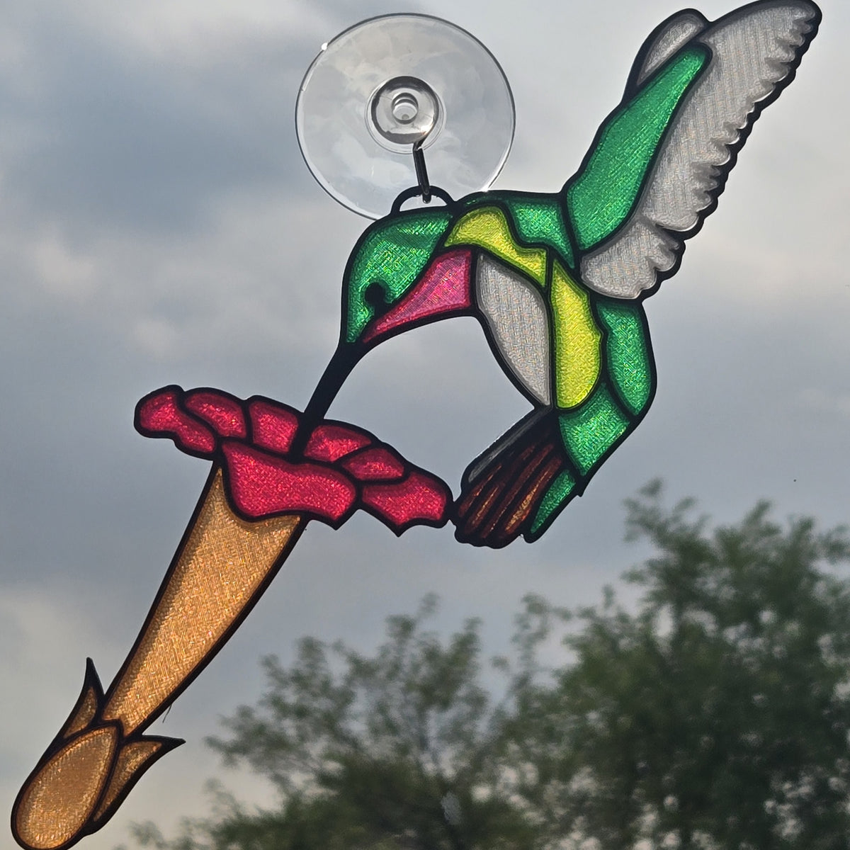 A 3D printed suncatcher depicting a green, yellow, and pink hummingbird perched on a red flower, suspended from a clear circular suction cup against an outdoor sky background.