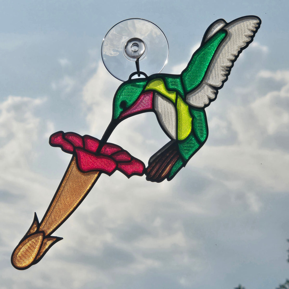A 3D printed suncatcher depicting a green and yellow hummingbird with pink wings, perched on a brown flower, attached to a clear suction cup, shown against a cloudy sky.