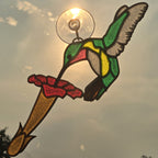 A 3D printed suncatcher shaped like a hummingbird feeding from a flower, mounted on a clear suction cup. The design features green, yellow, red, and brown glass panels with dark metal outlines, shown backlit by the sun against a cloudy sky.