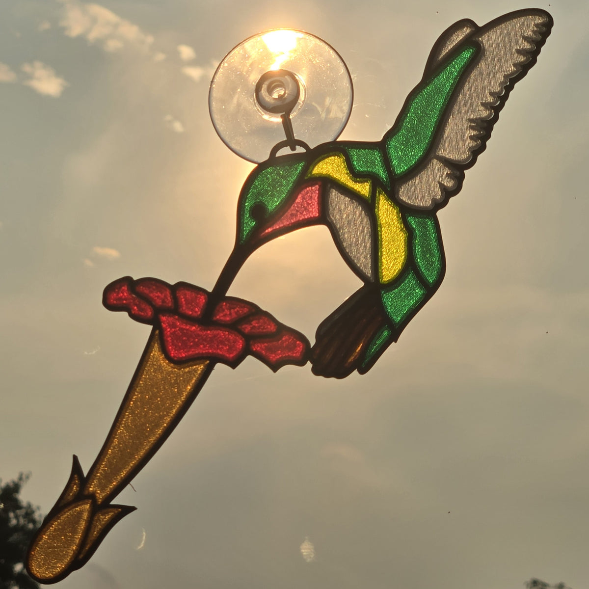A 3D printed suncatcher shaped like a hummingbird feeding from a flower, mounted on a clear suction cup. The design features green, yellow, red, and brown glass panels with dark metal outlines, shown backlit by the sun against a cloudy sky.